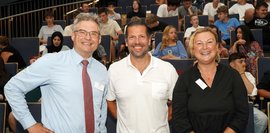 Zwei Männer und eine Frau, die für ein Bild posieren und im Hintergrund sitzen viele Schüler auf eine Tribühne