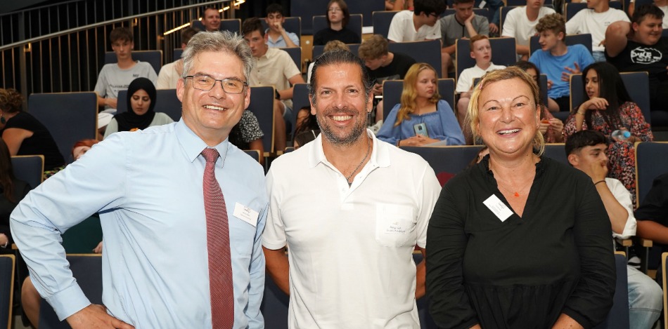 Abschlussveranstaltung 2023 Zwei Männer und eine Frau, die für ein Bild posieren und im Hintergrund sitzen viele Schüler auf eine Tribühne