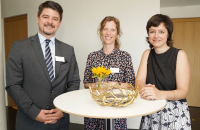 Ein Mann und zwei Frauen, die hinter einem Stehtisch stehen mit Blumen darauf und für ein Foto posieren