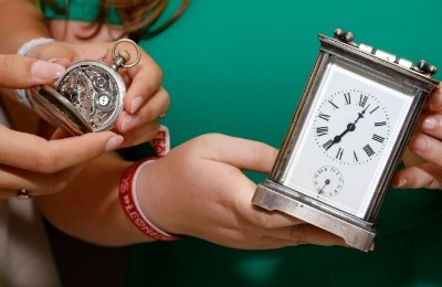 Eine kleine Wanduhr und eine Taschenuhr, die vor einer grünen Wand in den Händen gehalten werden